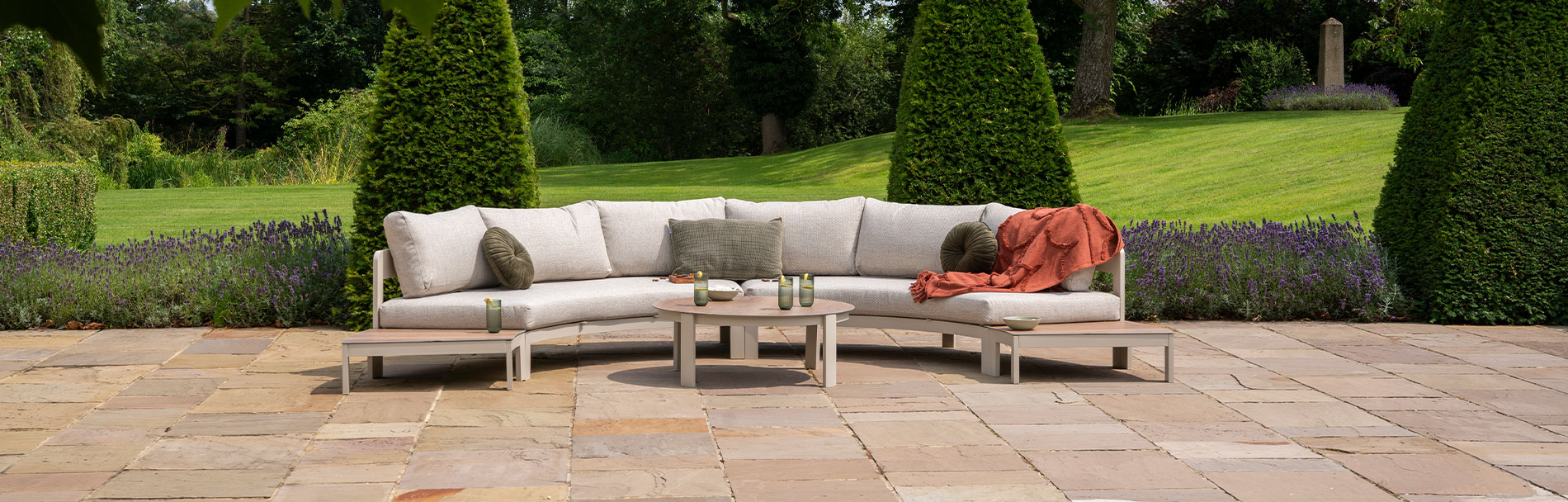 Millie Pebble curved corner garden sofa on a stone patio with a backdrop of trees